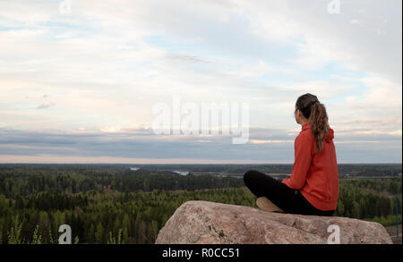 Young beautiful woman doing yoga and mindfulness exercises, while sitting on the edge of a hill and staring to the sunset horizon. Peaceful moment. Stock Photo