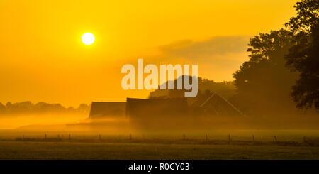 Orange sunrise over misty farmyard with barns and tractor in Twente, Netherlands Stock Photo