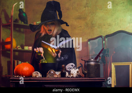 Portrait of witch in black hat reading spell over pot with green steam Stock Photo