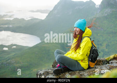 Image of happy tourist woman with backpack sitting on top of mountain in background of picturesque landscape Stock Photo