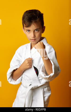 Little serious boy wearing white fighter kimono and standing in pose on yellow background Stock Photo