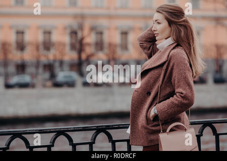 Bfashionable coat, beautiful woman dressed in stylish coat, keeps hand in pocket, focused into distance, has walk at city, thinks bout something, has  Stock Photo