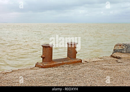 Steel rusty dual berth bitt on a fragment of old concrete pier on the background yellowish sandy seawater after the st Stock Photo