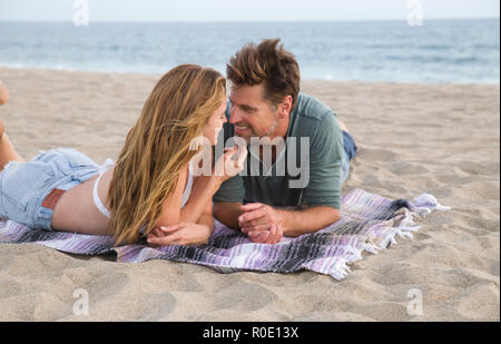 Smiling Mid-Adult Couple Looking at Each Other While Laying on Blanket at Beach Stock Photo