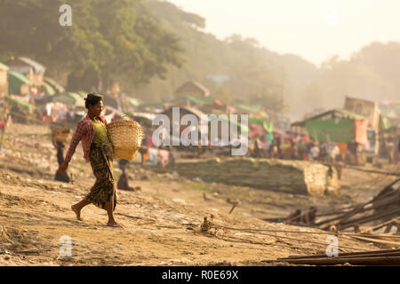 MANDALAY,MYANMAR,JANUARY 17, 2015 : A woman is carrying a basket , waling on a sloping ground and going to unload a boat of wooden planks in a slum ar Stock Photo