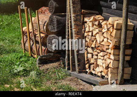 Stack of firewood next to old wooden cottage wall, sun lit grass lawn in front of it. Stock Photo