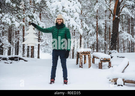 Full growth portrait of cheerful smiling male demonstrates white artificial fir tree, stands in winter forest has happy expression, glad to spend time Stock Photo