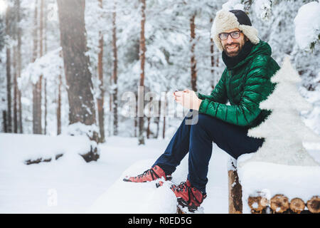 Photo of cheerful male types messages on smart phone looks directly into camera, sits in white winter forest, smiles happily, enjoys communication, wa Stock Photo