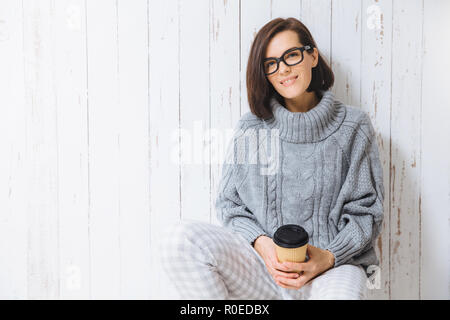 Adorable young woman wears warm sweater, spectacles, enjoys hot coffee or cappuccino from paper cup, sits on floor, being in high spirit, has coffee b Stock Photo