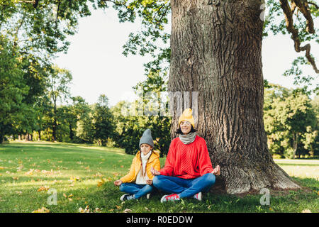 Yoga and meditation concept. Beautiful young woman in knitted clothes and small child keep eyes closed, meditate outdoors in green park, feel relaxed, Stock Photo