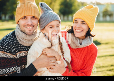 Playful little child with pigtails wears warm clothes, spends free time with lovely affectionate parents, have happy expression, feel relaxed. Three y Stock Photo