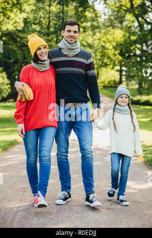 Portrait of friendly family walk across park. Beautiful brunette female in knitted yellow hat and sweater, handsome man embraces his wife and holds da Stock Photo