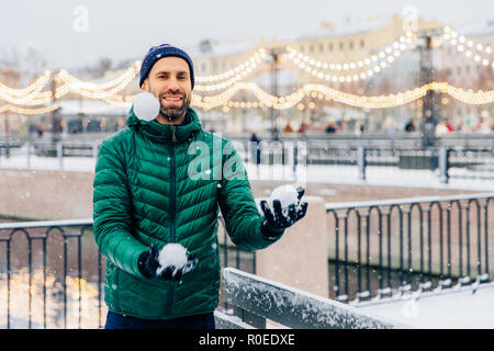 Playful smiling male juggles with snowballs throws them in air, has happy expression, stands against beautiful winter backgrounds. Happy overjoyed mal Stock Photo