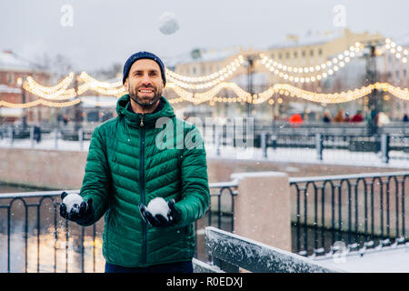 Pleasant looking male model keeps snowballs in hands going to have battle, waits for friends, looks happily into distance, being in good mood, stands  Stock Photo