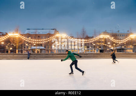 Active sorty male has fun in outdoor park on ice rink decorated with garlands, shows his talents of skating, makes fast movements on skates, being con Stock Photo