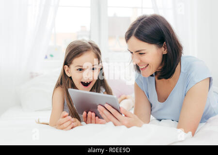 Beautiful mother and daughter watch something funny on tablet computer, connected to wireless internet, spend free time in bedroom, have surprised hap Stock Photo
