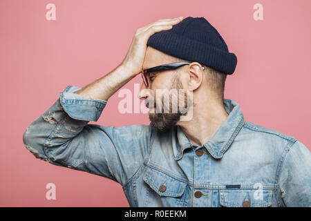 Sideways portrait of bearded male has regretful expression, keeps hand on forehead, looks desperately, isolated over pink background. Stressful unshav Stock Photo