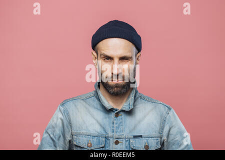 Photo of good looking bearded male blinks eye and looks seriously into camera, wears denim jacket and black hat, isolated over pink background. Stylis Stock Photo