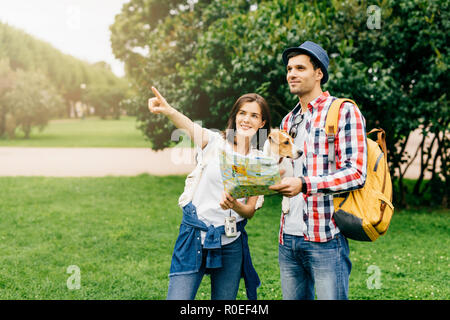 Young man traveller with his dog wearing hat and checkered shirt, losting in city, asking for way in passerby who is indicating with her forefinger on Stock Photo