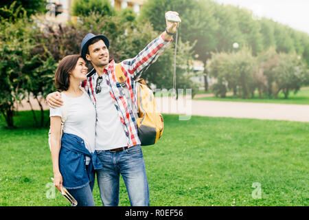 Romantic couple in love wearing casual clothes while standing on green grass, embracing each other and making selfie, smiling pleasanttly into camera. Stock Photo