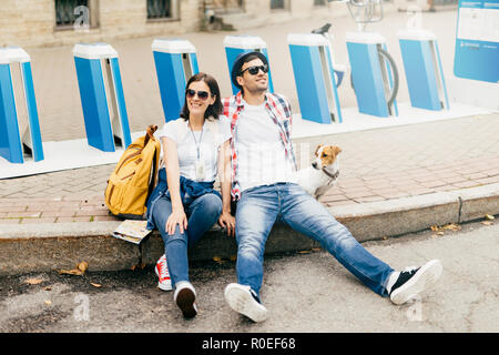 Carefree tourists sitting on pavement being tired after long excursion across city, being pleased to see many places of interest. Stylish male and fem Stock Photo