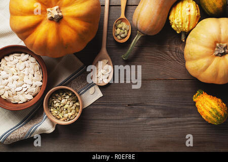 Frame of pumpkins of different sizes on a wooden background. A plate and a bowl of pumpkin seeds are standing on a kitchen towel. Two spoons with seed Stock Photo