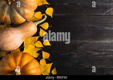 Three pumpkins of different sizes on a black wooden background with yellow leaves. Located on one side. Copy space. Top view. Stock Photo