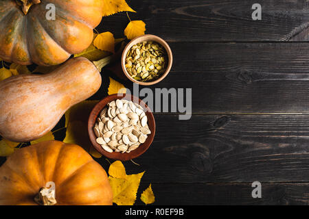 Three pumpkins of different sizes on a black wooden background with yellow leaves. Ceramic plate and wooden bowl with pumpkin seeds. Located on one si Stock Photo