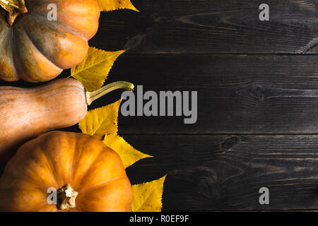 Three pumpkins of different sizes on a black wooden background with yellow leaves. Located on one side. Copy space. Top view. Stock Photo