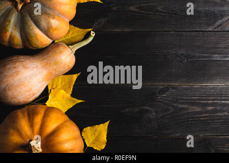 Three pumpkins of different sizes on a black wooden background with yellow leaves. Located on one side. Copy space. Top view. Stock Photo