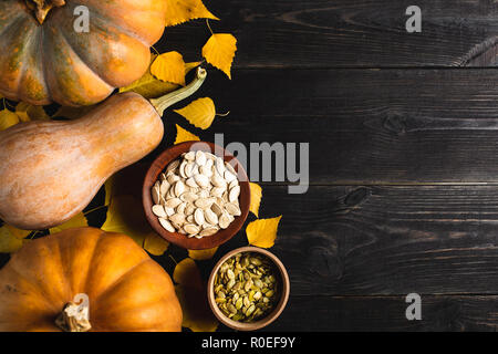 Three pumpkins of different sizes on a black wooden background with yellow leaves. Ceramic plate and wooden bowl with pumpkin seeds. Located on one si Stock Photo