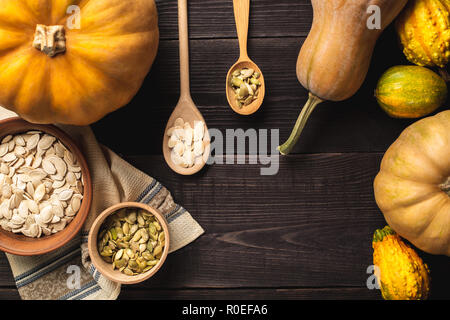 Frame of pumpkins of different sizes on a wooden background. A plate and a bowl of pumpkin seeds are standing on a kitchen towel. Two spoons with seed Stock Photo