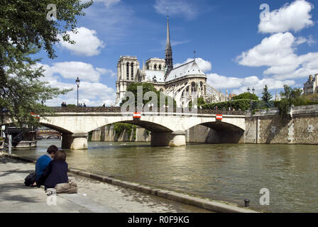 A couple sits on the Seine river embankment next to the Pont de l'Archeveche (Archbishop's Bridge) and Notre Dame cathedral in Paris, France. Stock Photo