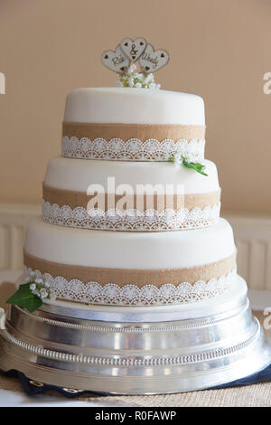 Three tier wedding cake in white icing with hessian trim and white lace, on a silver cake stand with a cream coloured wall background Stock Photo