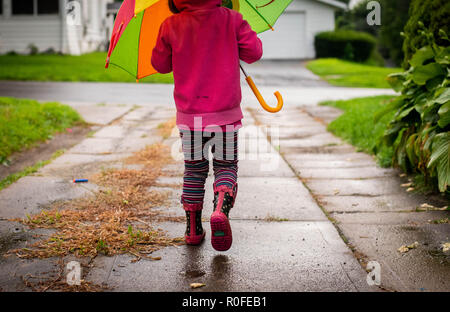 A 4-year old girl walks in the rain holding an umbrella and wearing rain boots. Stock Photo