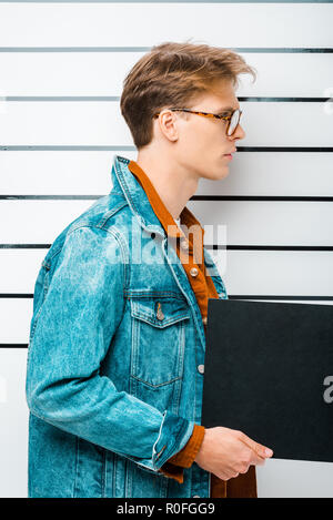 side view of arrested hipster man in eyeglasses holding empty prison board in front of police line up Stock Photo