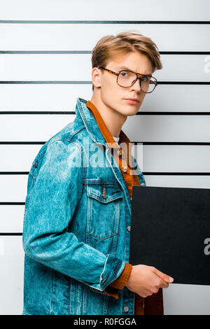 arrested hipster man in eyeglasses holding empty prison board and looking at camera in front of police line up Stock Photo