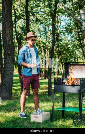 african american man in sunglasses and hat setting fire on grill in park Stock Photo