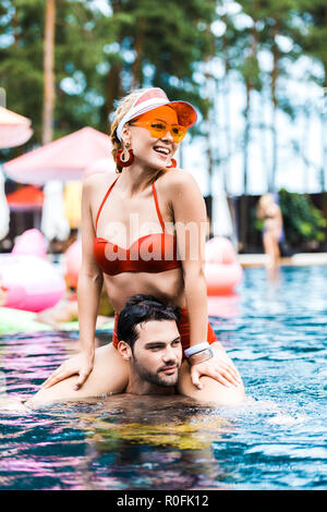 smiling woman in red swimsuit looking away while sitting on boyfriends shoulders in swimming pool Stock Photo