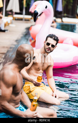 two male friends in sunglasses talking to each other and drinking beer on poolside Stock Photo