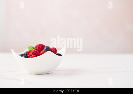 Fresh blueberries and raspberries with mint leaf in white bowl on white marble table over rose background with copy space. Stock Photo