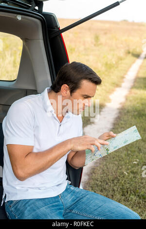 attractive man sitting in car trunk and navigating with map in field Stock Photo