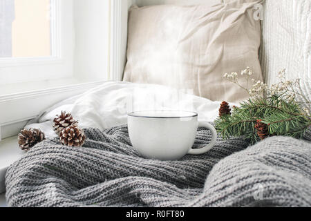 Cozy winter morning breakfast in bed still life scene. Steaming cup of hot coffee, tea standing near window. Christmas concept. Pillows, Pine cones and fir tree branch on wool plaid. Stock Photo