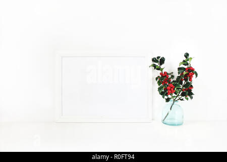 Horizontal white blank wooden frame mockup. Holly berry branches in blue glass vase on white table. Styled stock feminine photography. Home decor. Christmas winter concept. Stock Photo
