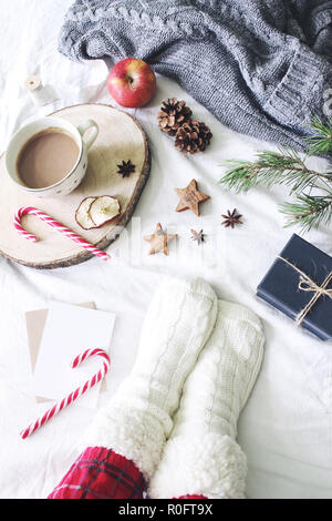 Christmas bed still life. Female legs in checkered pyjamas and woolen socks. Cup of coffee, candy canes and gift box. White linen bed sheet background with sweater. Winter flatlay, top view. Stock Photo