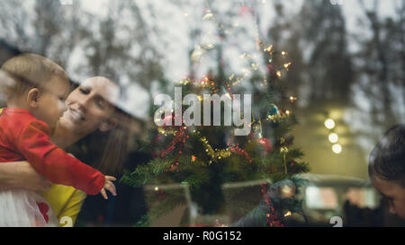 Retro image of a happy young mother holding her baby girl admiring Christmas tree which is still being decorated by her toddler son. View through a wi Stock Photo