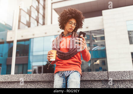 A handsome curly-haired guy of mixed race is dressed in a trendy youth-style scarf and sweater writes a message in a teleone against the background of Stock Photo