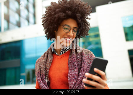 A handsome curly-haired guy of mixed race is dressed in a trendy youth-style scarf and sweater writes a message in a teleone against the background of Stock Photo