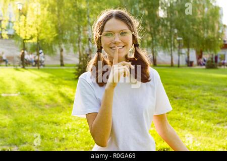 Outdoor portrait een girl shows sign quietly, secret, holds finger near her lips. Background city park at sunset. Stock Photo