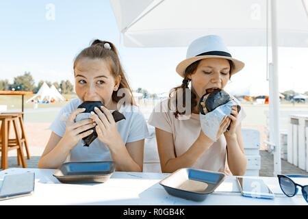 Teens girls with an appetite eats black fast food burger. Summer street cafe, recreation area, city park background. Stock Photo